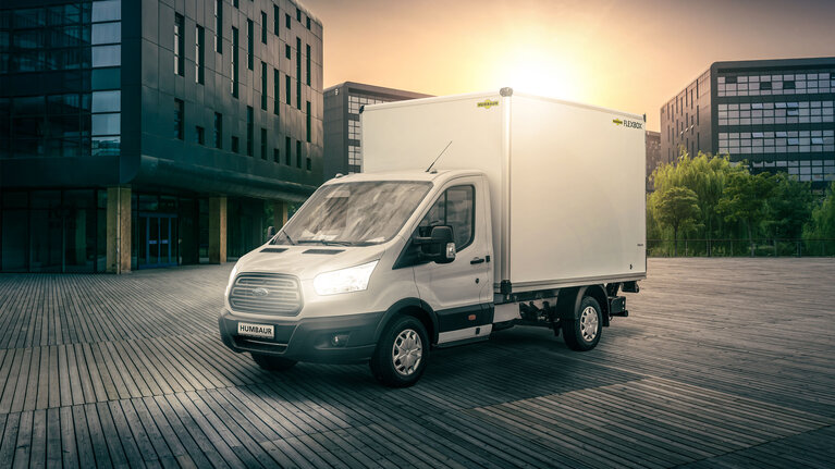 Photograph of a white Ford van with a white Humbaur Flexbox in front of an urban backdrop.